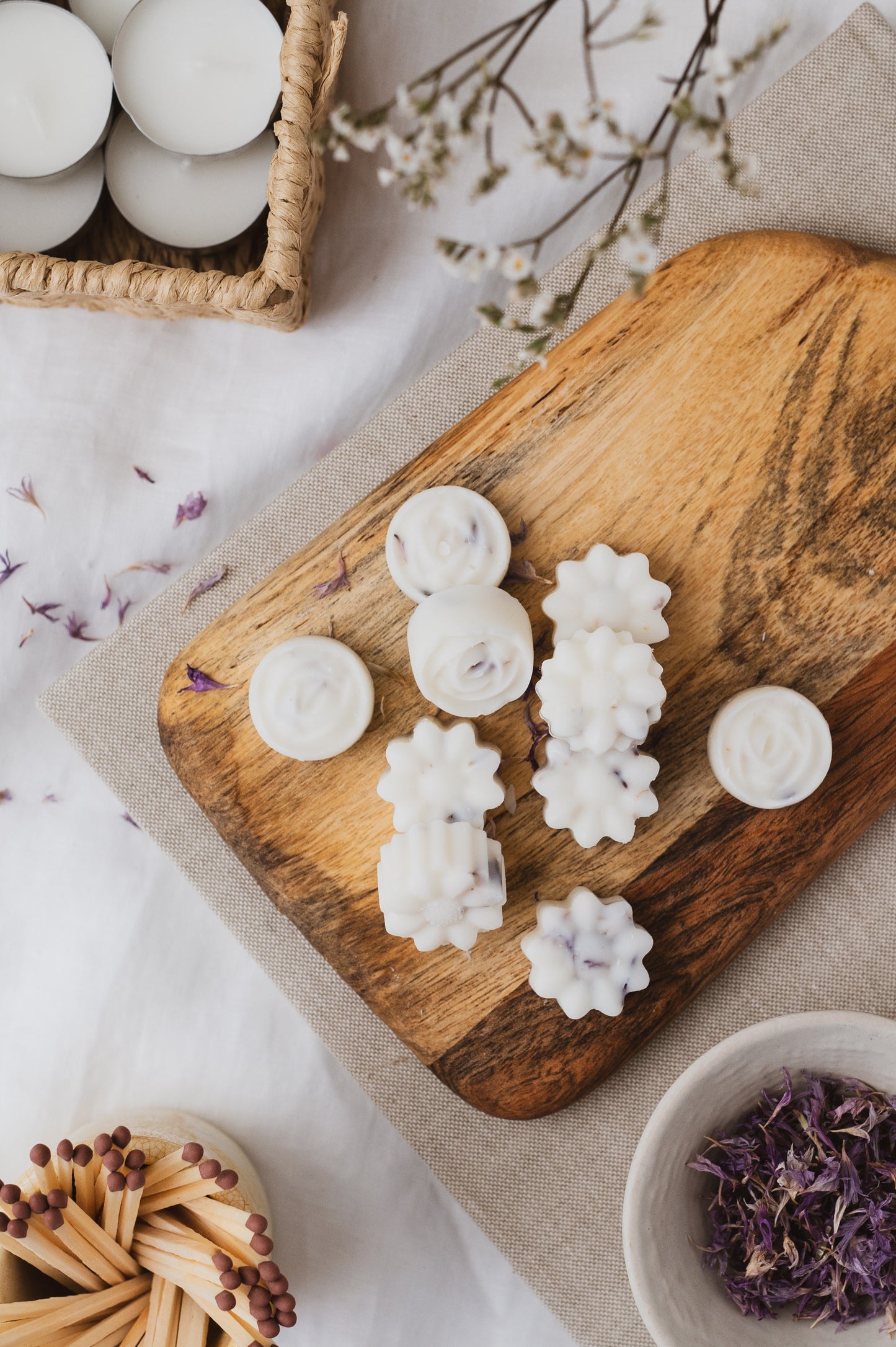 Flower-shaped botanical soy wax melts resting on a wooden chopping board. Matches, plant, petals and tealights for decoration.